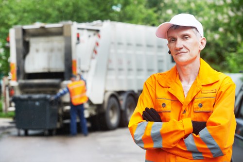 A professional rubbish removal worker wearing an orange high-visibility jacket with reflective silver stripes and a white cap is standing outdoors on a paved area, with arms crossed and a neutral expression. In the background, a large white waste disposal truck is parked, with its rear open and a worker in a high-visibility vest actively loading or managing waste materials. The environment includes green foliage and trees, indicating a scene in a suburban or urban area near Cricklewood. The scene depicts a typical rubbish collection or clearance operation, with the focus on the worker in the foreground and the waste vehicle behind, illustrating the process of garden or property clearance in the local area, as part of services offered by Garden Clearance Cricklewood.
