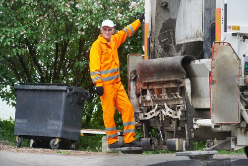 A waste collection worker dressed in bright orange high-visibility safety clothing, including a jacket and trousers with reflective strips, standing next to a large industrial rubbish compactor or garbage truck on a paved street. The worker is smiling and appears to be operating or inspecting the machinery, with one foot resting on the truck's platform. To the left, there is a black wheeled waste bin positioned on the curb, near green leafy bushes and trees providing a natural backdrop. The scene is well-lit with natural daylight, emphasizing the clean, functional environment typical of rubbish removal operations in a suburban or urban setting, possibly within the Cricklewood area. The image showcases the practical aspect of rubbish collection services offered by Garden Clearance Cricklewood, highlighting the professional handling of waste materials and the use of specialized equipment for effective rubbish disposal, set against a typical residential or commercial exterior environment.