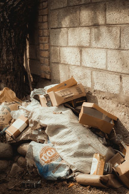 A large accumulation of black rubbish bags and discarded items such as cardboard and plastic packaging is piled in front of a red metal door embedded in a concrete wall. The door has graffiti tags, a 