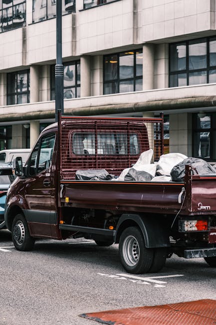 A small maroon flatbed truck parked on a city street in front of a modern building with large glass windows and concrete facade. The truck's cargo area is filled with various types of waste materials 